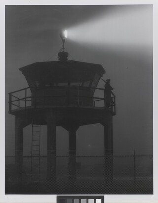 A black-and-white photograph of a person standing in silhouette atop a tower with a bright spotlight shining from the top.