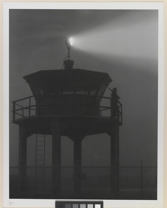 A black-and-white photograph of a person standing in silhouette atop a tower with a bright spotlight shining from the top.