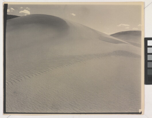 A sepia-toned photograph of a rippling sand dune rising up into a sky dotted with a few small clouds.