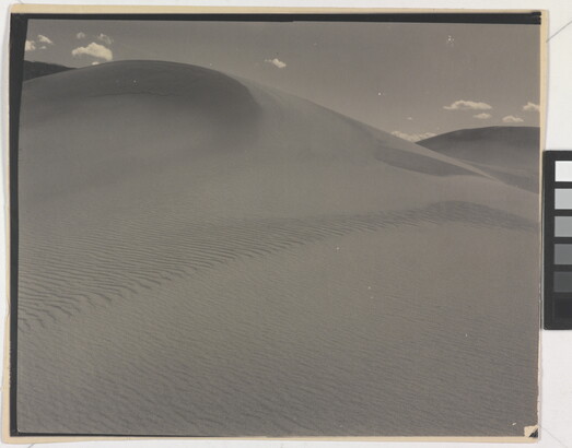 A sepia-toned photograph of a rippling sand dune rising up into a sky dotted with a few small clouds.