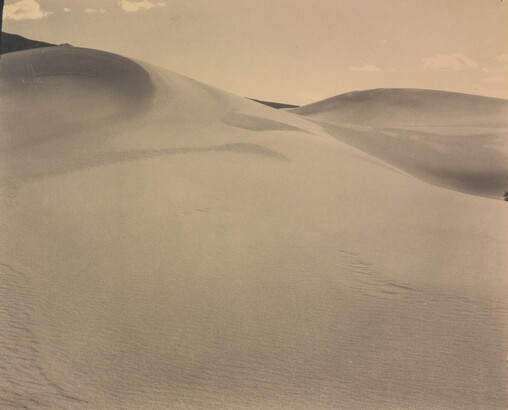 A sepia-toned photograph of a rippling sand dune rising up into a sky dotted with a few small clouds.