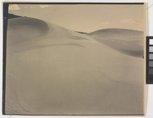 A sepia-toned photograph of a rippling sand dune rising up into a sky dotted with a few small clouds.