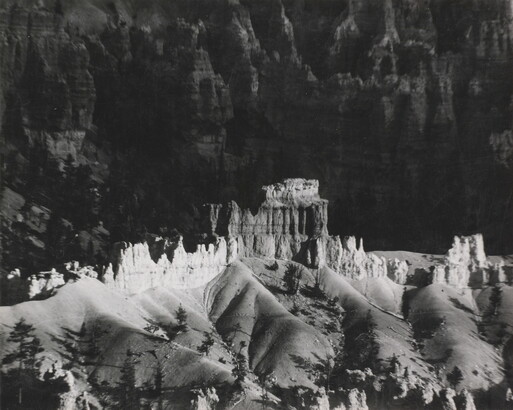 A black-and-white photograph of a landscape with hoodoos, or vertical spires of rock, on top of sand dune shapes with cliffs in the background.