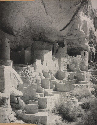 A black-and-white photograph of an abandoned, crumbling rock city built into the side of a desert cliff.