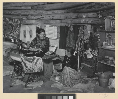 A black-and-white photograph of two Indigenous women seated, talking, and sewing inside of a low-ceilinged, wood structure.