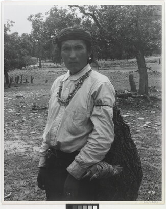 A black-and-white photograph of an Indigenous man carrying a large bag.