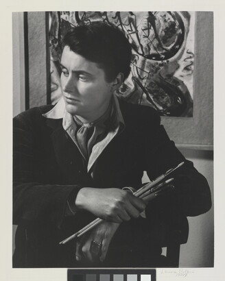 A black-and-white portrait photograph of a seated White woman holding paintbrushes in front of an abstract painting.