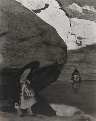 A black-and-white photograph of a woman with a long headscarf standing at the edge of a water hole surrounded by boulders and rock walls.