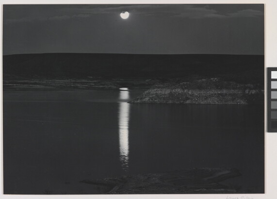 A black-and-white photograph of a night sky and full moon reflected across calm water.
