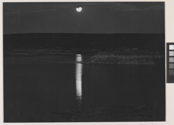 A black-and-white photograph of a night sky and full moon reflected across calm water.