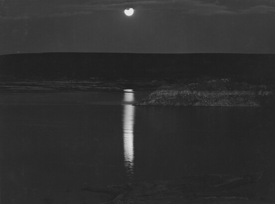 A black-and-white photograph of a night sky and full moon reflected across calm water.