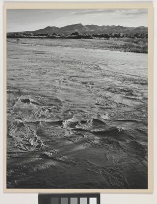 A black-and-white photograph of a choppy river and then grassy plains and mountains beyond the shore.