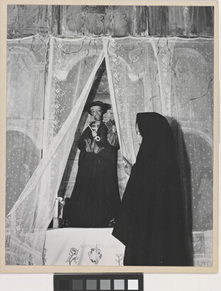 A black-and-white photograph of a woman wearing a black shawl over her head facing a statue of a saint holding a large key.
