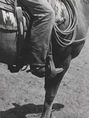 A black-and-white photograph of a person&#039;s leg in a cowboy boot, including spurs, sitting astride a horse, viewing only the horse&#039;s front quarter.