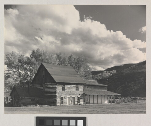 A black-and-white photograph of a three-story log building with mountains and large puffy clouds in the background.