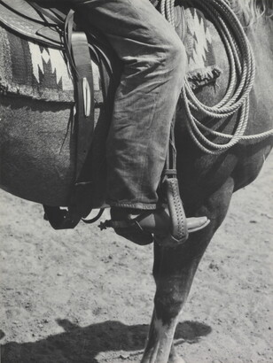 A black-and-white photograph of a person&#039;s leg in a cowboy boot, including spurs, sitting astride a horse, viewing only the horse&#039;s front quarter.
