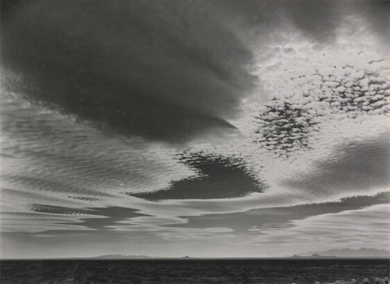 A black-and-white photograph of an expansive desert plain under rippled clouds with mountains in the far distance.