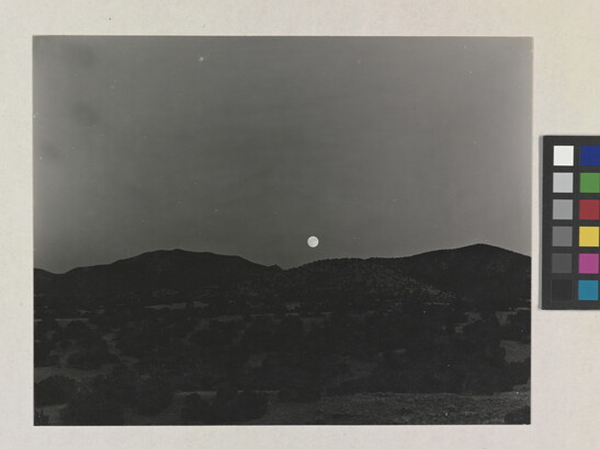 A black-and-white photograph of the moon rising above a mountain landscape.