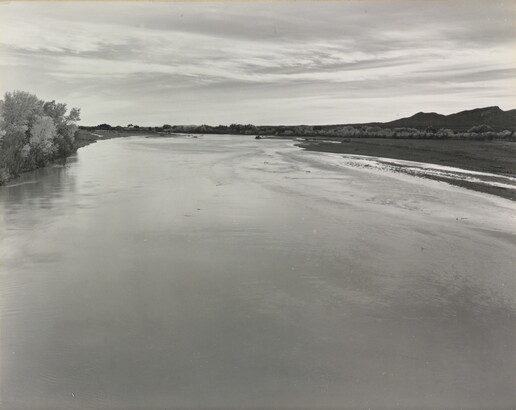 A black-and-white photograph of a calm river with trees on the left shore and mountains in the distance.