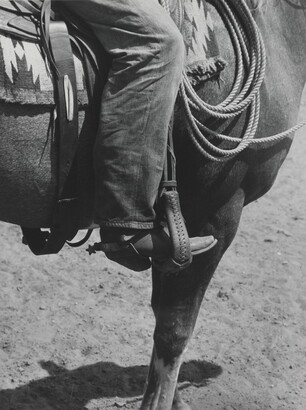 A black-and-white photograph of a person&#039;s leg in a cowboy boot, including spurs, sitting astride a horse, viewing only the horse&#039;s front quarter.