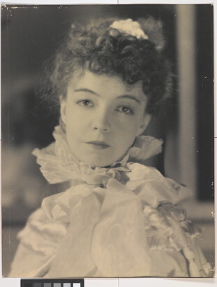 A black-and-white portrait photograph of a White woman with curly hair pulled on top of her head, wearing a top with a ruffled collar.