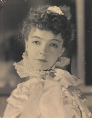 A black-and-white portrait photograph of a White woman with curly hair pulled on top of her head, wearing a top with a ruffled collar.