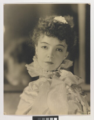 A black-and-white portrait photograph of a White woman with curly hair pulled on top of her head, wearing a top with a ruffled collar.