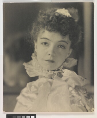 A black-and-white portrait photograph of a White woman with curly hair pulled on top of her head, wearing a top with a ruffled collar.