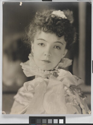 A black-and-white portrait photograph of a White woman with curly hair pulled on top of her head, wearing a top with a ruffled collar.