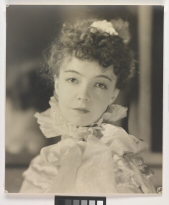A black-and-white portrait photograph of a White woman with curly hair pulled on top of her head, wearing a top with a ruffled collar.