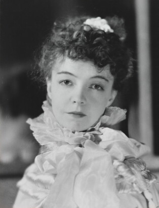 A black-and-white portrait photograph of a White woman with curly hair pulled on top of her head, wearing a top with a ruffled collar.