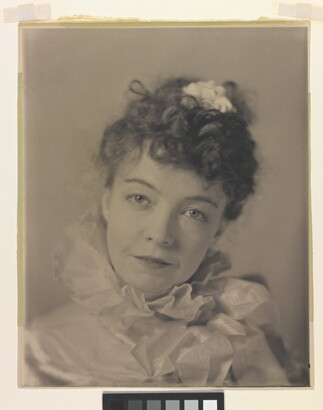 A black-and-white portrait photograph of a White woman with curly hair pulled on top of her head, wearing a top with a ruffled collar.