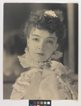 A black-and-white portrait photograph of a White woman with curly hair pulled on top of her head, wearing a top with a ruffled collar.