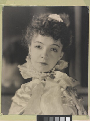 A black-and-white portrait photograph of a White woman with curly hair pulled on top of her head, wearing a top with a ruffled collar.