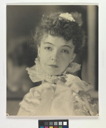 A black-and-white portrait photograph of a White woman with curly hair pulled on top of her head, wearing a top with a ruffled collar.