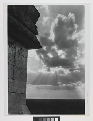 A black-and-white photograph of the sun&#039;s rays bursting from clouds and a stark brick architectural feature in the left foreground.