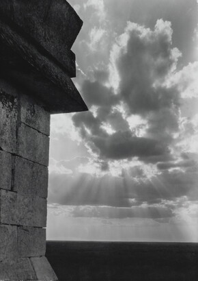 A black-and-white photograph of the sun&#039;s rays bursting from clouds and a stark brick architectural feature in the left foreground.