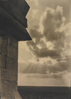 A sepia-toned photograph of the sun&#039;s rays bursting from clouds and a stark brick architectural feature in the left foreground.