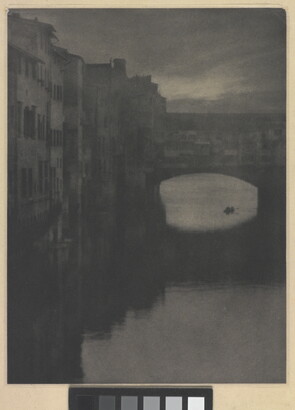 A sepia-toned photograph of a wall of tall buildings lining a river and an enclosed arched bridge stretching over the water.