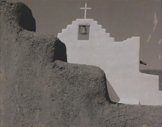 A black-and-white photograph of a terraced adobe bell tower with a cross on top, partially hidden by a terraced adobe wall.