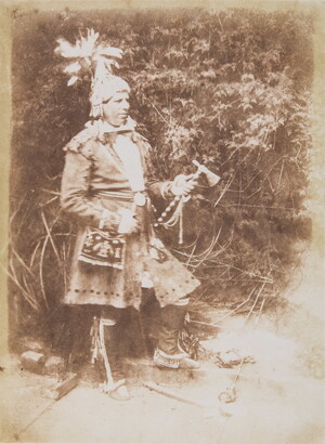 A sepia-toned photograph of an Indigenous man wearing traditional clothing and holding a tomahawk.