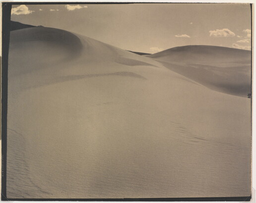 A sepia-toned photograph of a rippling sand dune rising up into a sky dotted with a few small clouds.