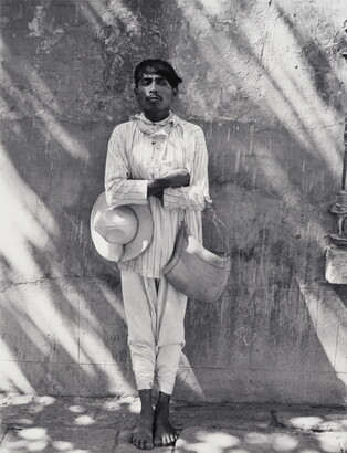 A black-and-white photograph of a dark-skinned man in light-colored shirt and pants, holding a hat under one crossed arm and a bag on the other, leaning against a wall.