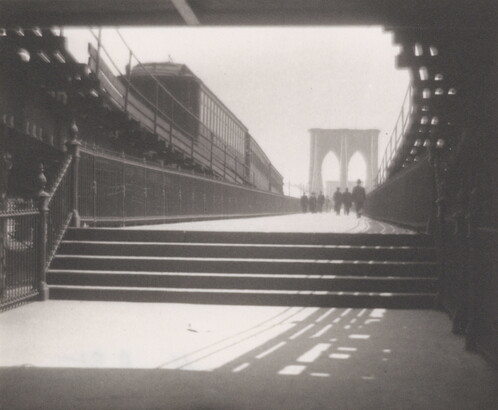 A black-and-white photograph of a set of stairs that leads to the tower of a suspension bridge and people walking toward the tower.