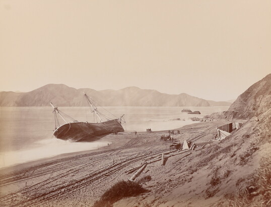 A black-and-white photograph of a large boat tipped on its side in the water near a sandy shore.