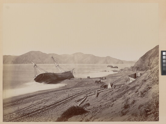 A black-and-white photograph of a large boat tipped on its side in the water near a sandy shore.