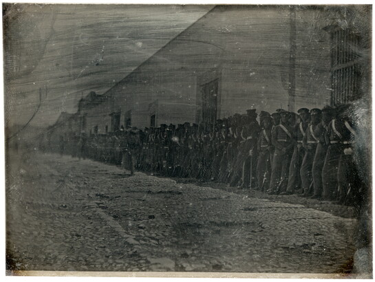 A dark, black-and-white photograph of a group of figures in military uniforms lined up against a building on a cobblestone street.