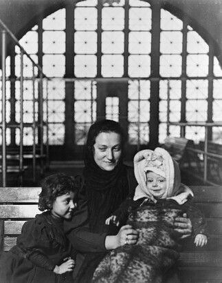 A black-and-white photograph of a White woman with two young children seated on a bench, with a large arched window behind them.