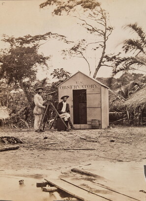 A black-and-white photograph of two men with a telescope standing in front of a small shack with &quot;U.S. Observatory&quot; above the doorway.