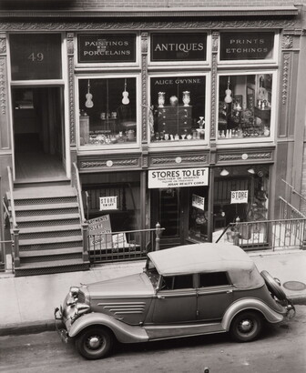 A black-and-white photograph of a vintage car parked on the street outside of a storefront advertising antiques.
